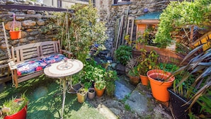 Un espace extérieur accueillant, aménagé avec un banc en bois et une table ronde, entouré de différents pots de plantes et d’un mur en pierre.