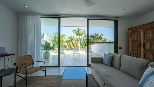Una sala moderna con sofá, una silla, una mesa de centro y un ventanal corredero que da a un balcón con vistas a palmeras.