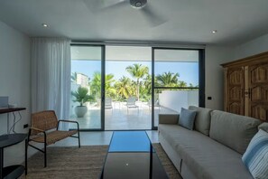 Una sala moderna con sofá, una silla, una mesa de centro y un ventanal corredero que da a un balcón con vistas a palmeras.