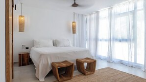 Un dormitorio moderno con una cama grande, dos mesitas de noche, un ventilador de techo y ventanas amplias con cortinas translúcidas.