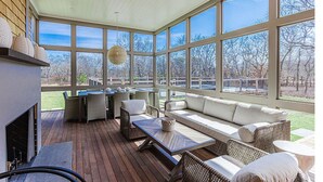 A sunlit patio with wicker furniture, a fireplace, and large windows offering a view of the outdoors. - Brand New Spacious Luxury Home on Edgartown Great Pond with Pool and Spa (Edgartown)