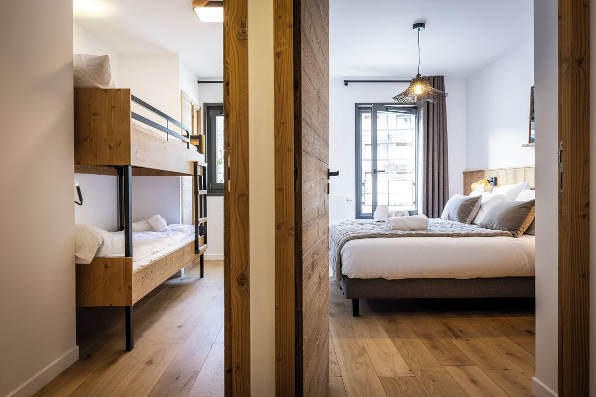 A room with a bunk bed, a single bed, wooden flooring, and a large window with curtains.