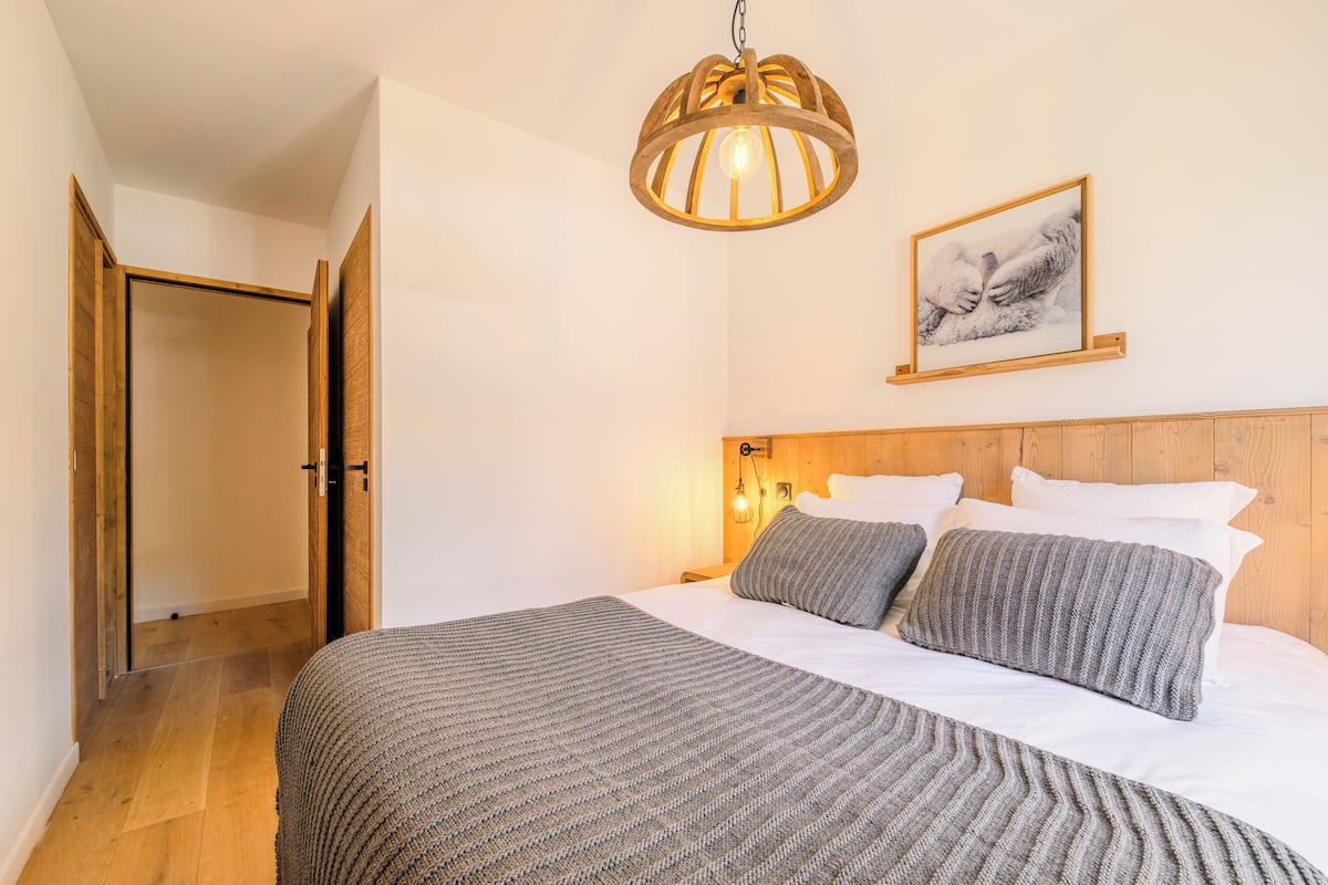 A bedroom with a wooden headboard, a large bed with grey bedding, a hanging light fixture, and a framed picture on the wall.