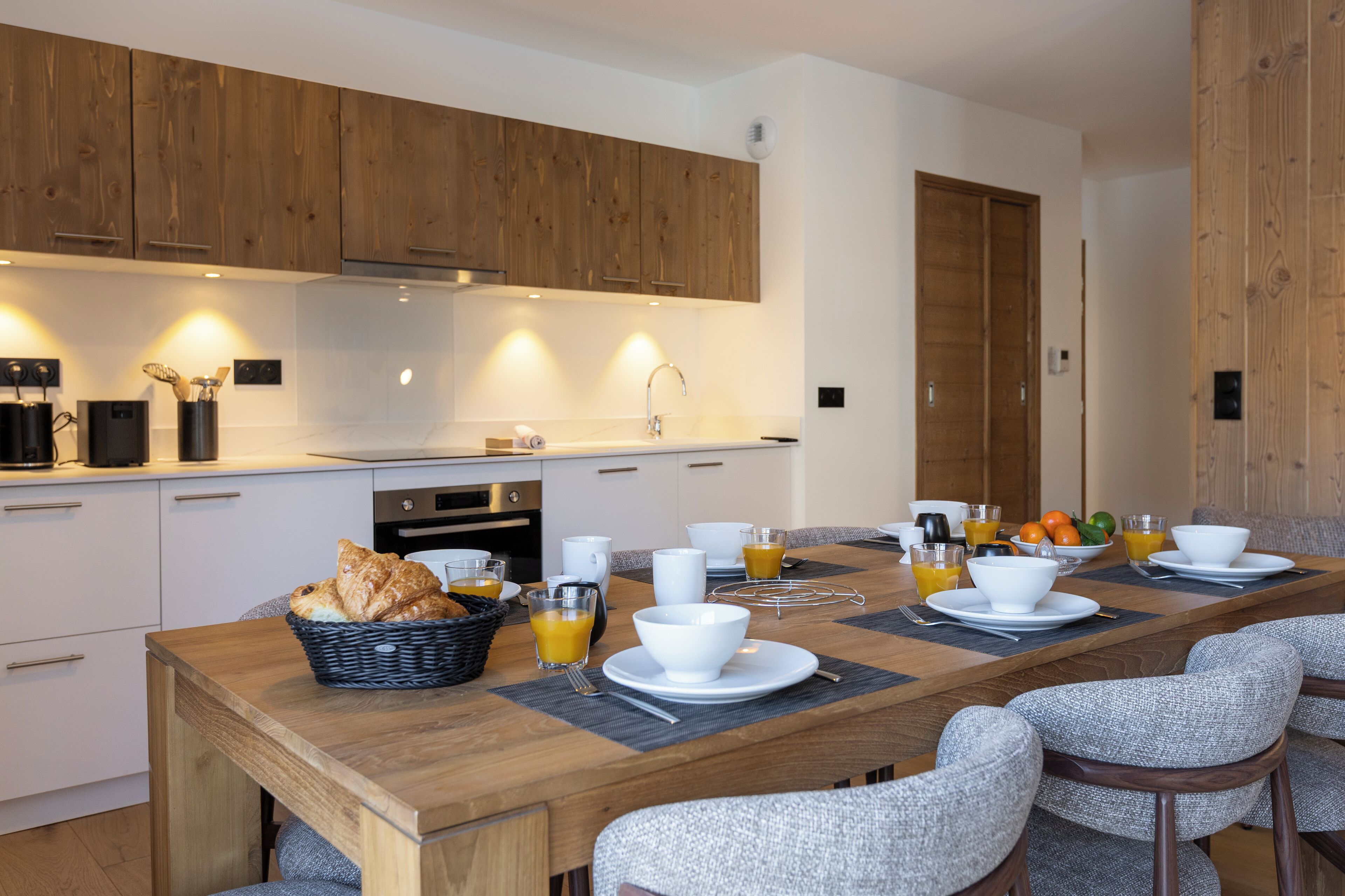 Eine moderne Küche mit Holzschränken, einem Esstisch, der für ein Frühstück gedeckt ist, mit Schüsseln, Tassen und einem Brotkorb.