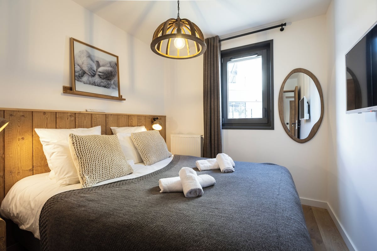 A neatly made bed with a gray blanket, two pillows, and two folded towels. There is a framed picture on the wall above the bed, a round mirror, and a television on the wall.