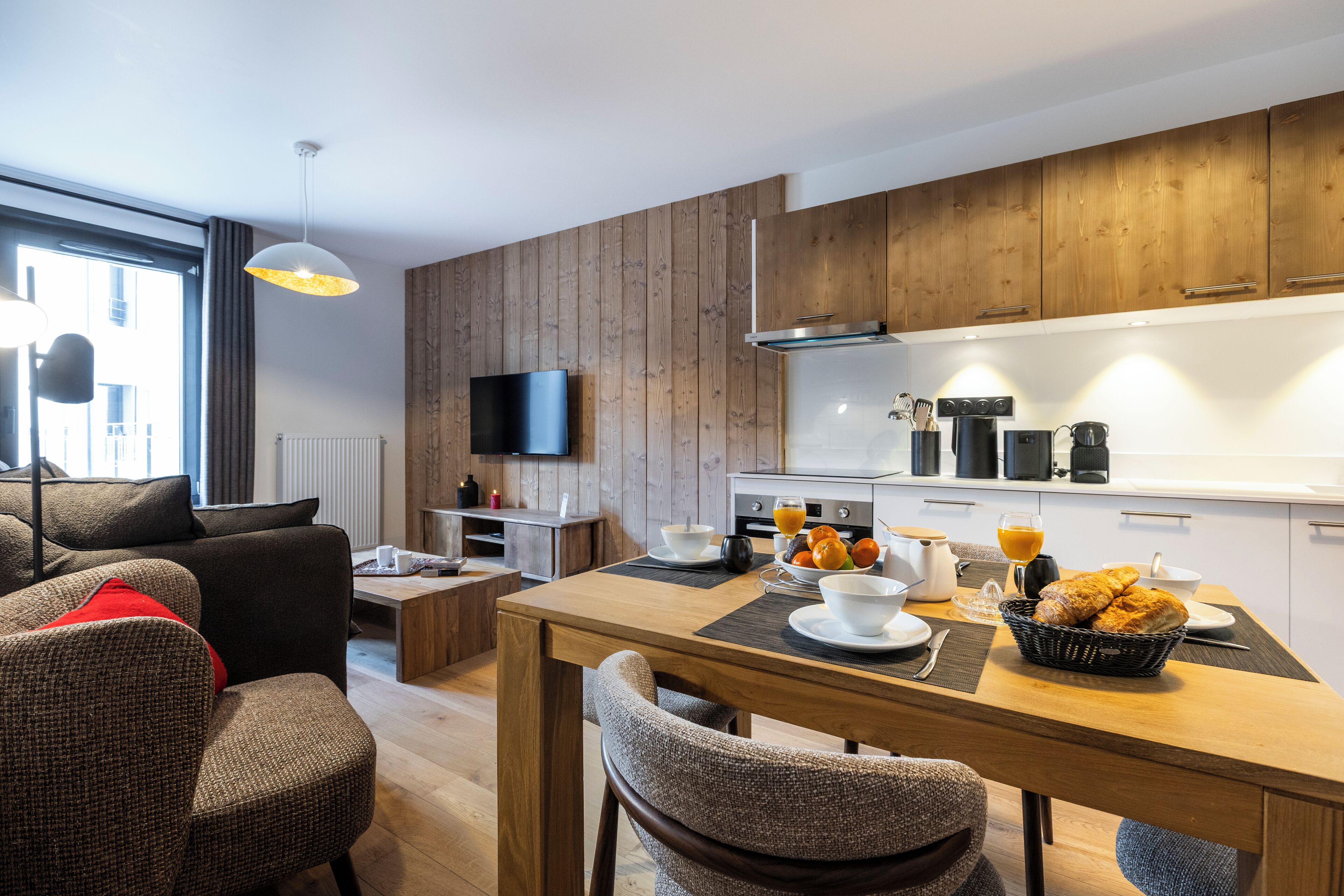 A modern living room with a sofa, a dining table with breakfast items, and a kitchen area with wooden cabinets and appliances.