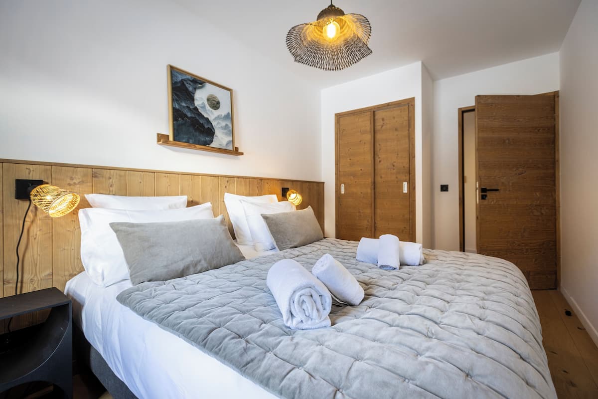 A neatly made bed with a grey quilt, white pillows, and rolled towels on a wooden headboard. There is a wall-mounted light fixture and a framed picture above the bed.