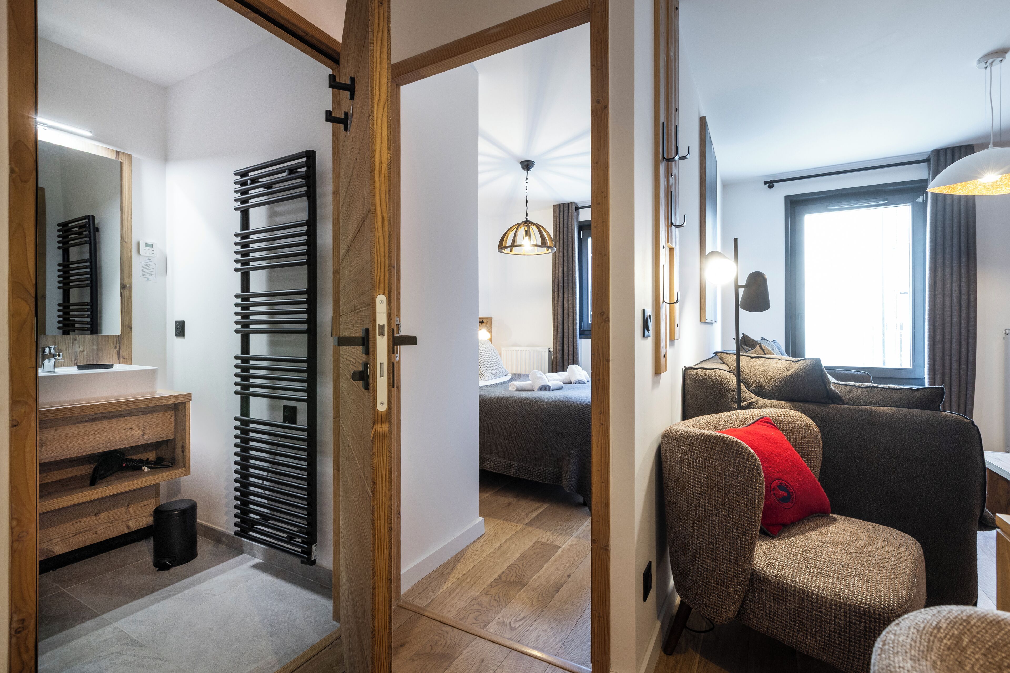 A modern hotel room with a wooden floor, a large bed, a sitting area with a red cushion, a large window, and a bathroom with a towel rack and sink.
