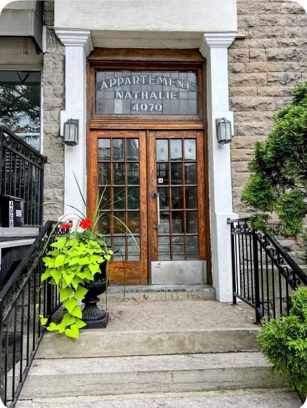 A stone building with a wooden double door, a sign reading 'APARTMENT NATHALIE 4070', and a potted plant on the steps.