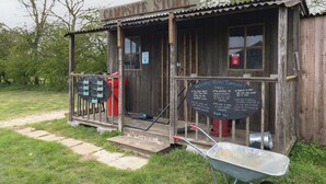 Una estructura de madera rústica con un letrero que dice 'Campsite Store' y una pizarra que anuncia los horarios de apertura y un mensaje de bienvenida.