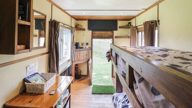 Un interior de cabaña acogedor, con paredes de madera, un escritorio pequeño con una cesta de libros, una ventana con cortinas y una litera con edredón estampado.