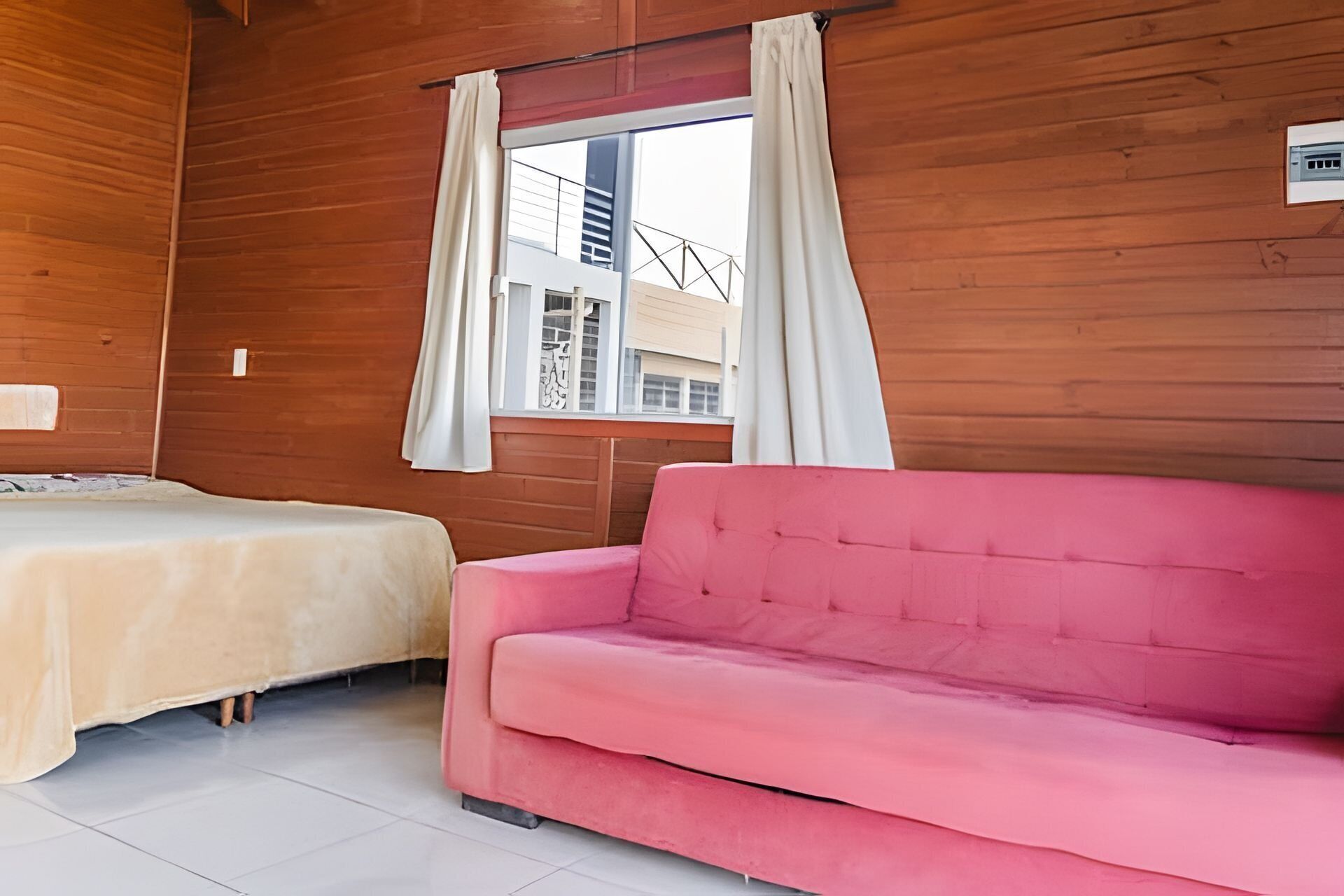 A room with two beds, a pink sofa, wooden walls, a window with curtains, and a view of buildings outside.