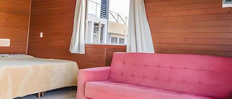 A room with two beds, a pink sofa, wooden walls, a window with curtains, and a view of buildings outside.