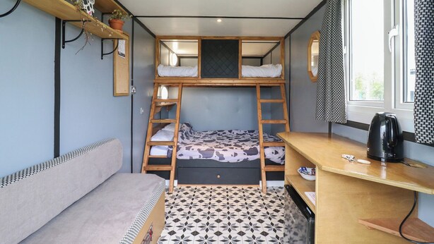 Ein kleines Zimmer mit Etagenbetten, einem Schreibtisch, einer Kaffeemaschine und einem Fenster mit gemusterten Vorhängen.
