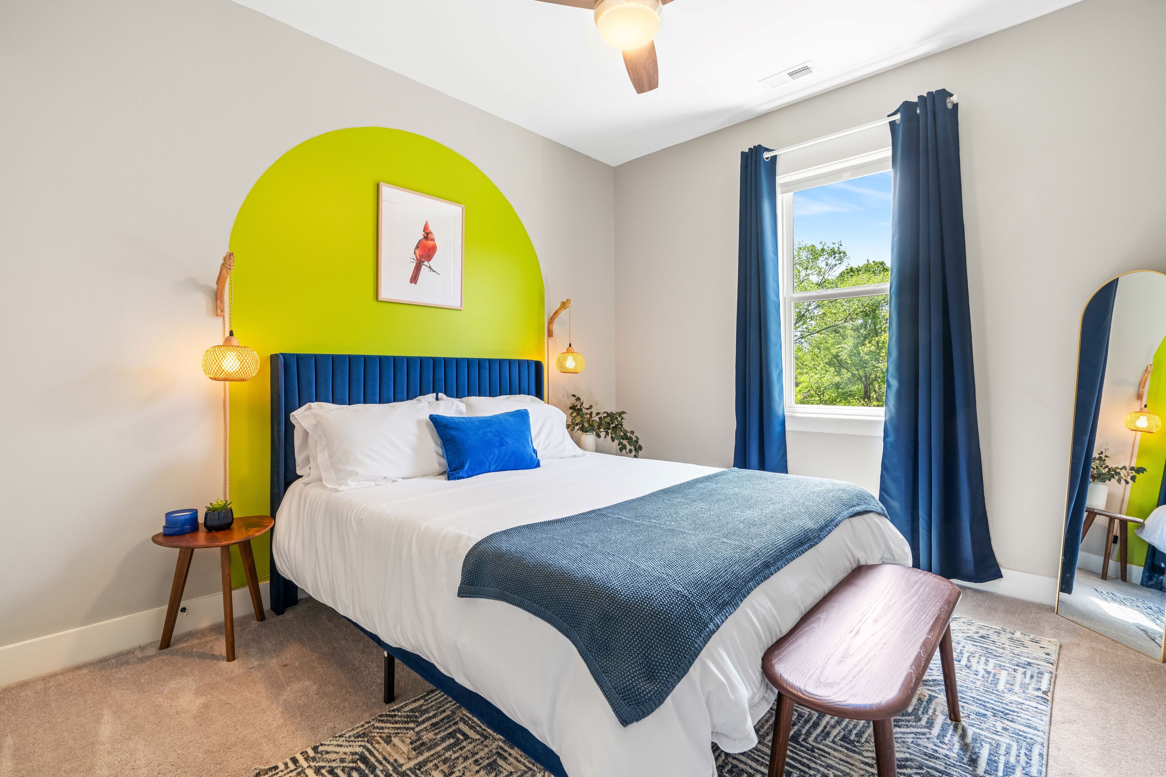 Un dormitorio con cama, un banco, una ventana con cortinas, un ventilador de techo y una lámpara de pared.