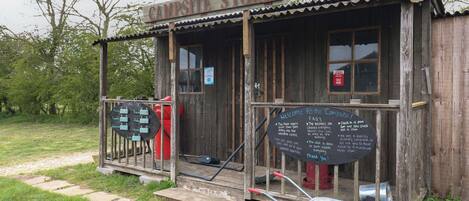 A rustic wooden structure labeled "Campsite Store" with a chalkboard sign listing opening times and a welcome message.