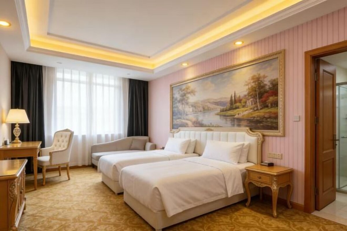 A hotel room with two beds, a desk, a chair, a painting on the wall, and a door.