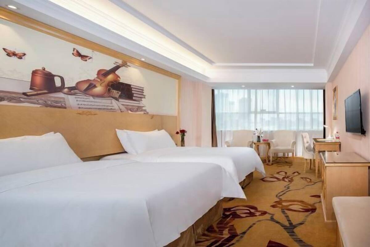 A hotel room with two beds, a TV, a desk, and a large window.