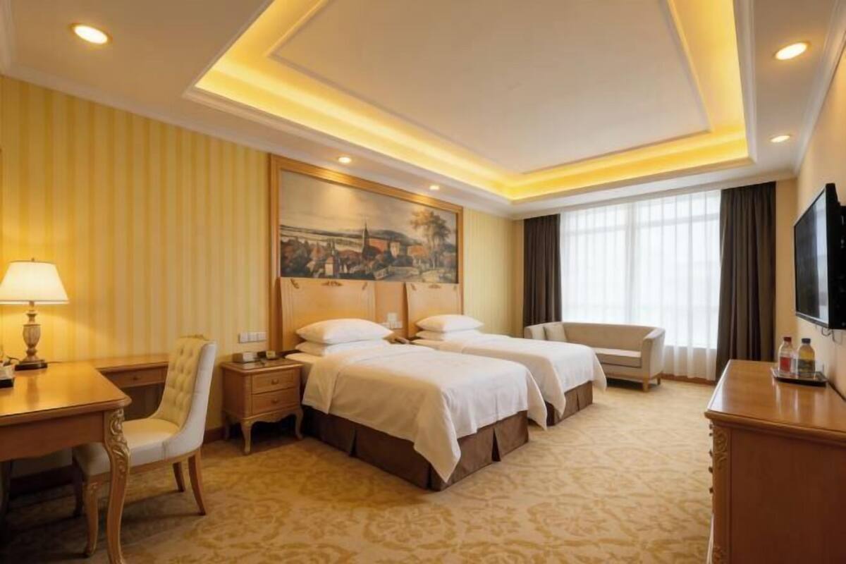 A hotel room with two beds, a desk, a chair, a TV, and a painting on the wall.