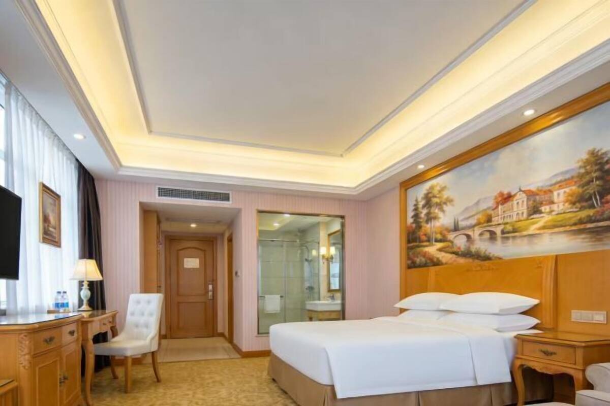 A hotel room with a bed, a desk, a chair, a painting on the wall, and a bathroom visible through a glass door.