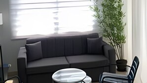 A modern living room with a grey sofa, a small round table, two chairs, a large window with blinds, and a potted plant. - City Center Apartment Torre Leonardo VI (Santo Domingo)