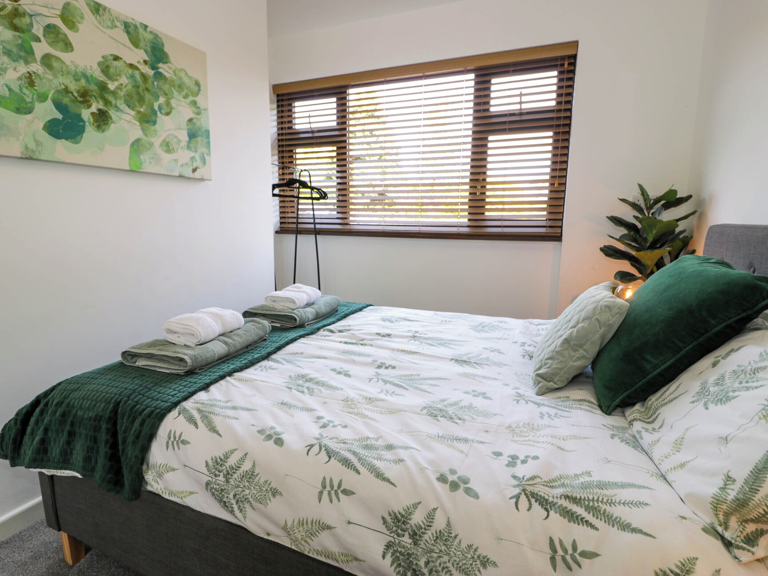 A neatly made bed with a green blanket and a white bedspread with a fern pattern. There are folded towels on the bed, a floor lamp, and a plant next to the bed.