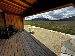 Een overdekt terras met houten banken en stoelen, een grindpad en uitzicht op een bergachtig landschap.