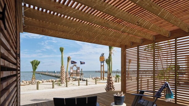 Eine Holzterrasse mit Blick auf den Strand, eine Liege und ein kleiner Tisch mit einem Getränk.