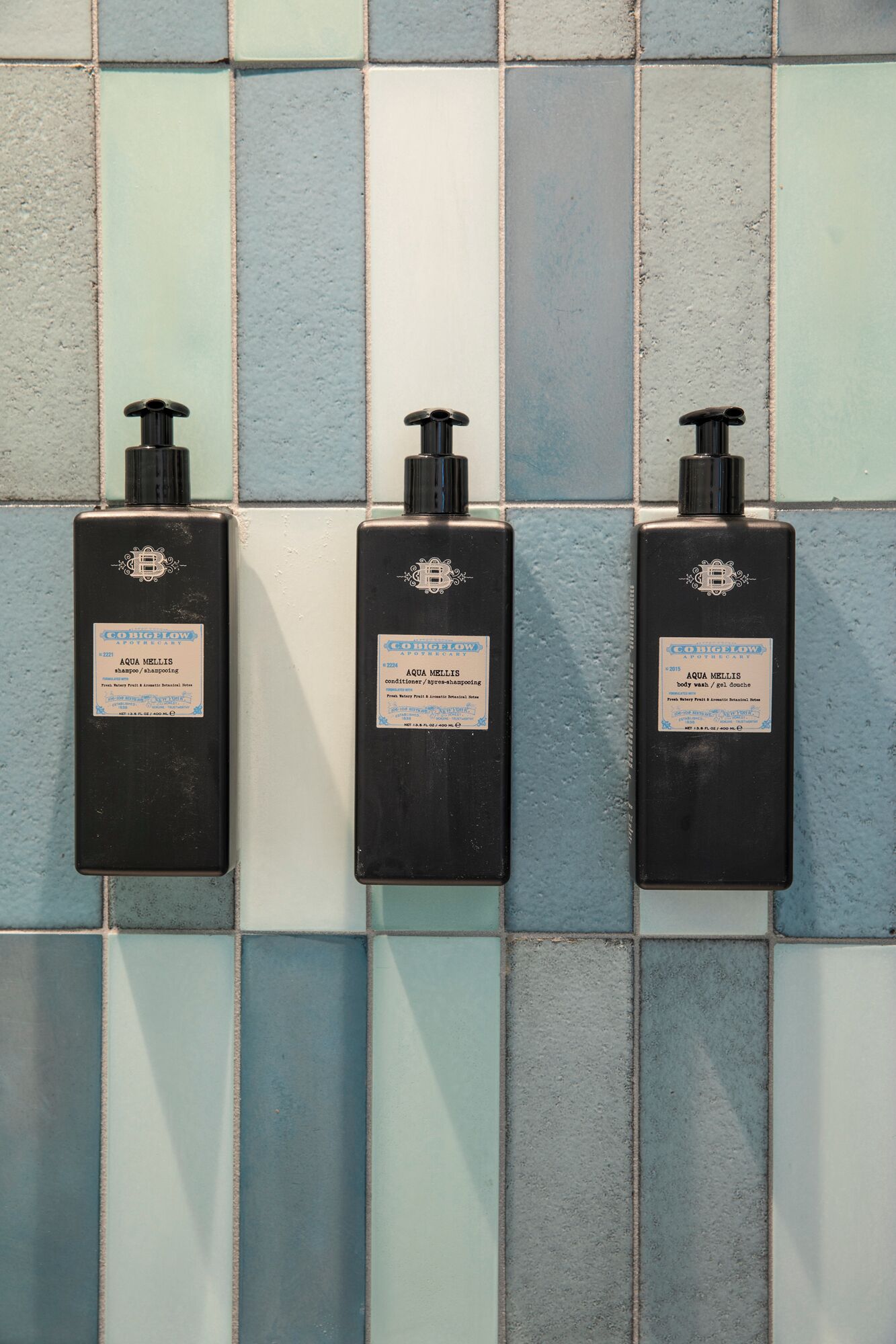 Trois bouteilles noires de gel douche et de shampoing Aqua Mellis sont posées sur un mur carrelé.