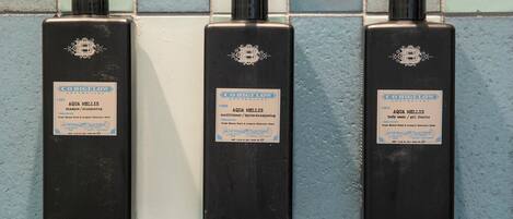 Trois bouteilles noires de gel douche et de shampoing Aqua Mellis sont posées sur un mur carrelé.