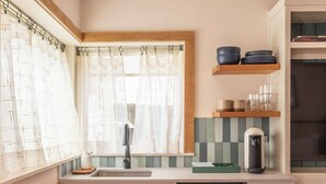 Eine kompakte Küche mit Geschirrspüler, Spüle und einem Fenster mit Vorhängen. Über der Arbeitsplatte befinden sich Regale mit Geschirr und einer Kaffeemaschine.