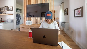 Eine Person benutzt einen Laptop an einem Holztisch in einer modernen Küche mit Holzelementen, einem Essbereich und einer Küchenarbeitsplatte mit verschiedenen Gegenständen.