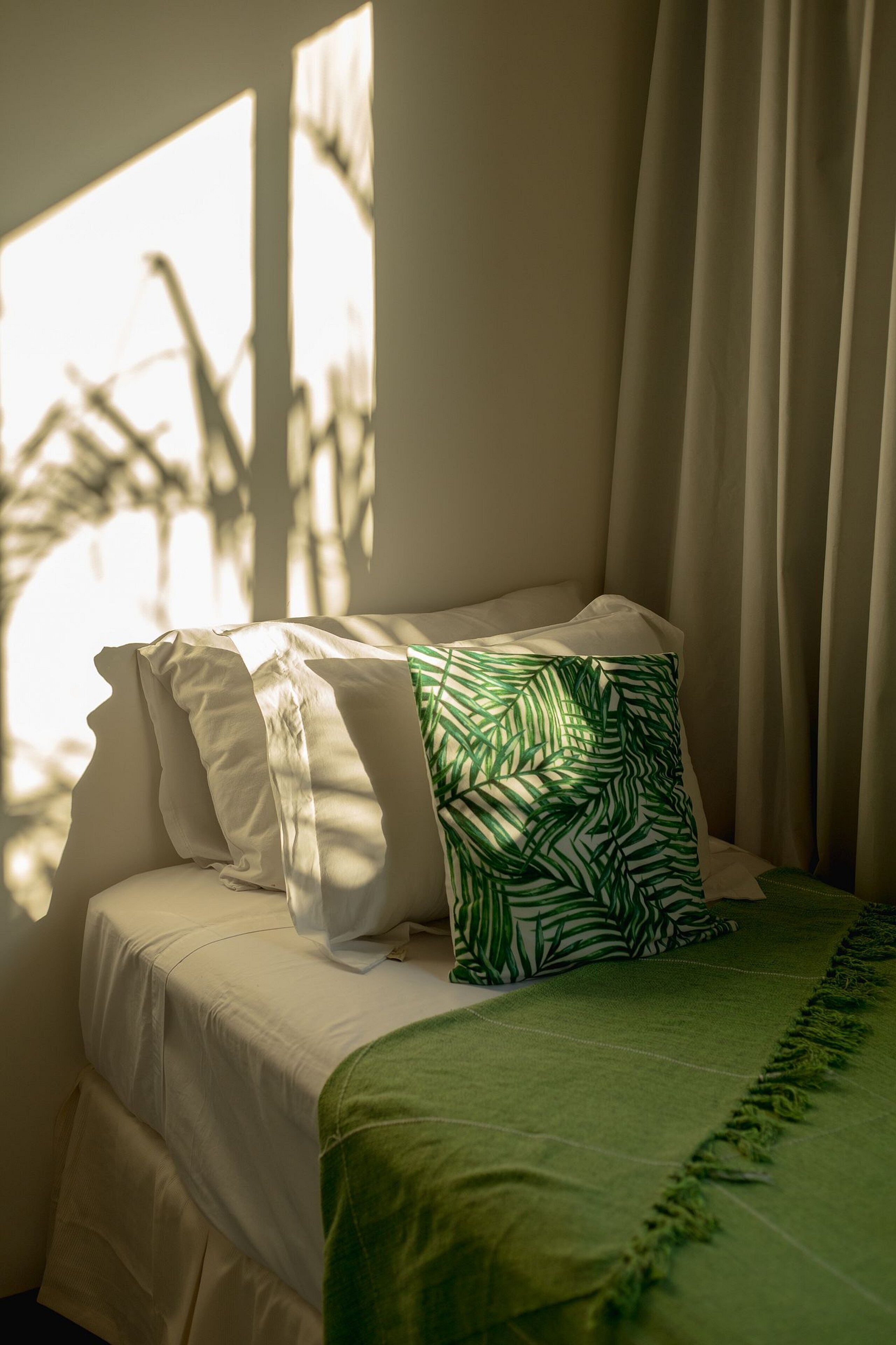 Un lit avec une literie blanche et un coussin orné d’un motif de feuilles vertes, placé près d’une fenêtre avec des rideaux légers.