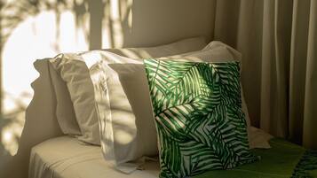 Un lit avec une literie blanche et un coussin orné d’un motif de feuilles vertes, placé près d’une fenêtre avec des rideaux légers.