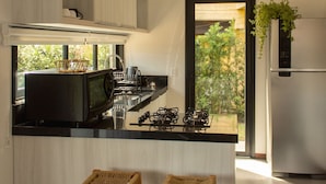 A modern kitchen with a central island, two wicker stools, a stainless steel refrigerator, and hanging pendant lights. - Casas Valley Boutique - Praia do Rosa (Imbituba)