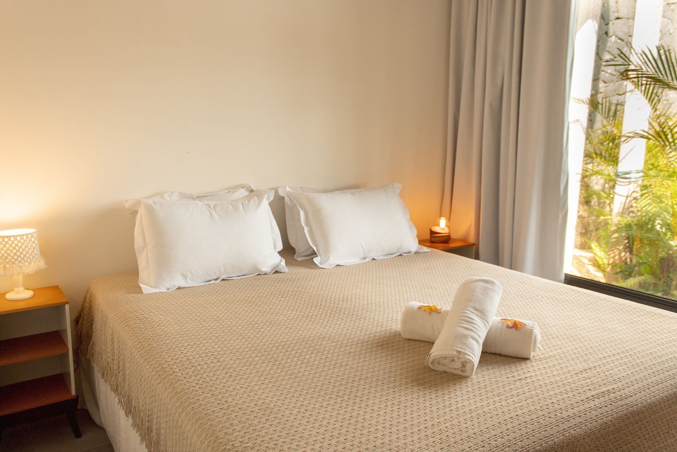 Uma cama bem arrumada com um edredom bege e duas almofadas, uma mesinha de cabeceira com um abajur, e uma janela com cortinas.