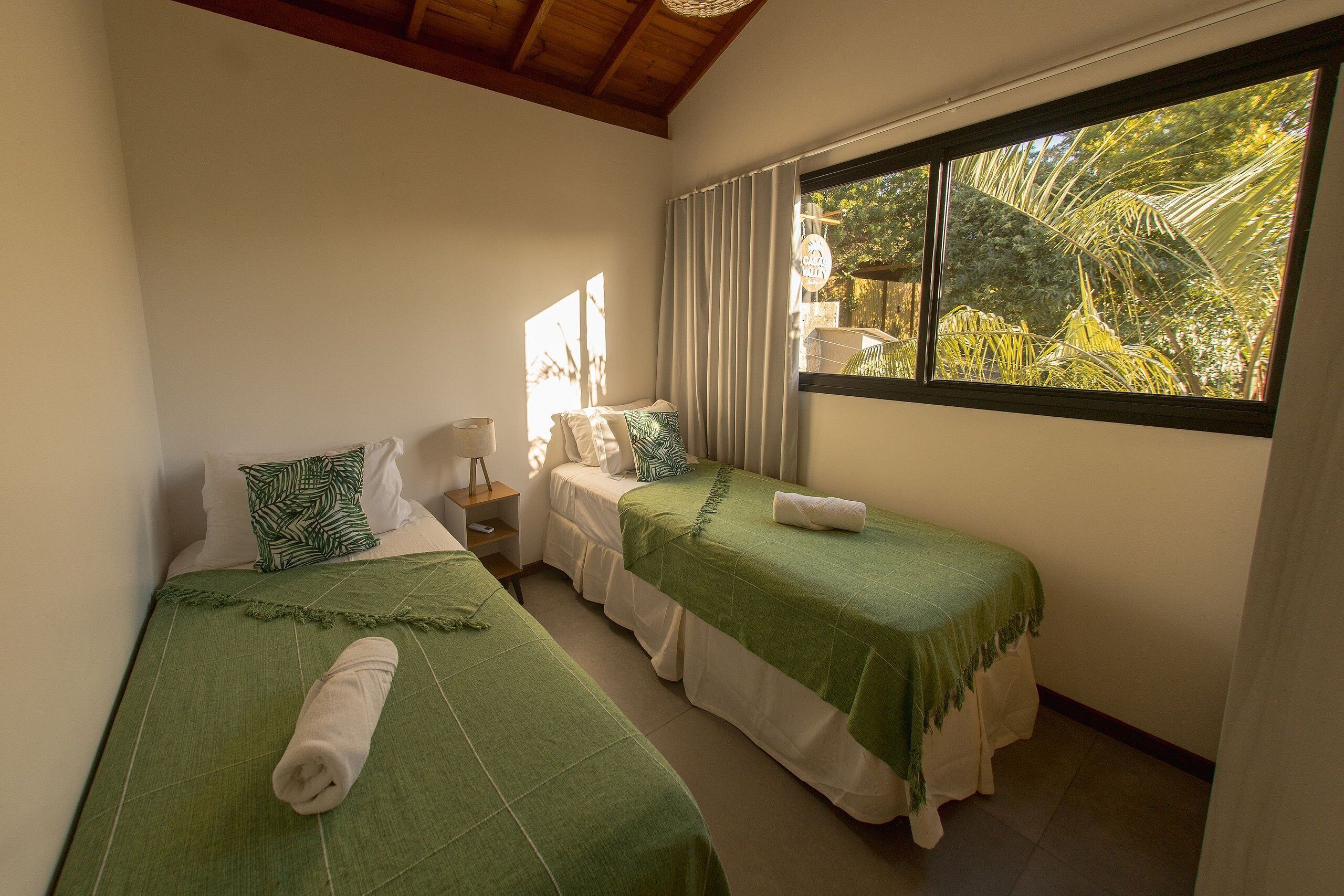 A room with two beds, each with a green bedspread and white pillows. There is a window with curtains, and a small table with a lamp.