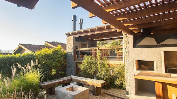A wooden deck with a stone fire pit and seating area, surrounded by greenery. - Casas Valley Boutique - Praia do Rosa (Imbituba)