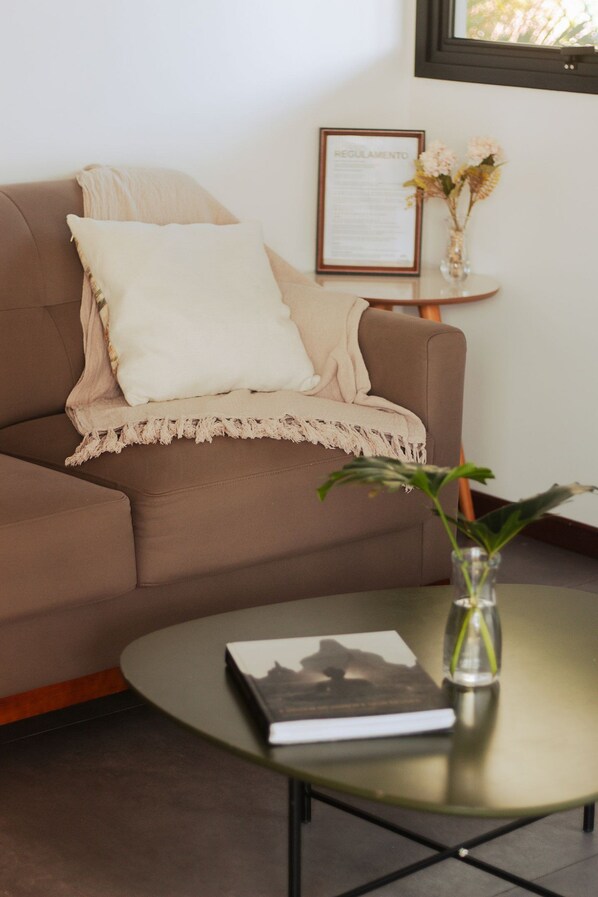 Un canapé marron avec un coussin blanc et un plaid beige. Un document encadré est posé sur une petite table à côté du canapé. Un vase rempli de fleurs se trouve sur la table.