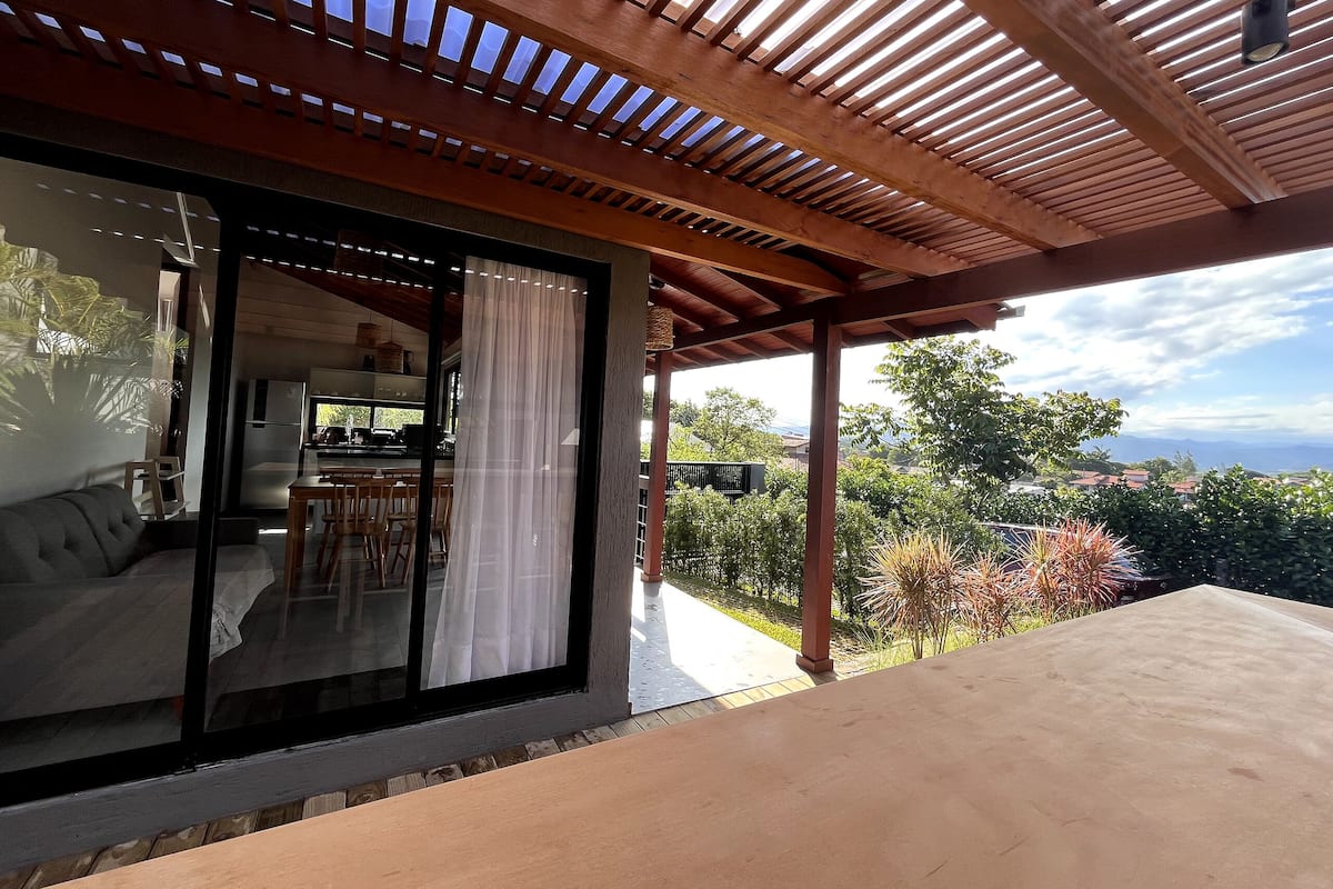 A covered outdoor area with a view of a kitchen and dining area, wooden ceiling beams, and a table.
