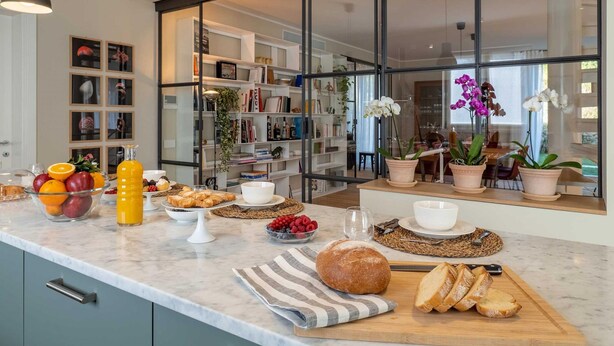 A modern kitchen with a marble countertop, a variety of fruits, pastries, and bread, along with a large window offering a view of the outdoors. - Luxury Golf Villa in Bogogno (Bogogno)