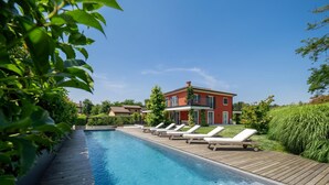A swimming pool with lounge chairs, surrounded by greenery and a red building in the background. - Luxury Golf Villa in Bogogno (Bogogno)