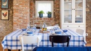 A dining area with a checkered tablecloth, white chairs, a white cabinet with glass doors, and a stone wall. - Green Lodge - Apartment 1 - 1 BR 1 Bath (Cressa)