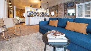 A modern living room with a blue sofa, a small table with a book, and a kitchen in the background. - Green Lodge - Apartment 1 - 1 BR 1 Bath (Cressa)