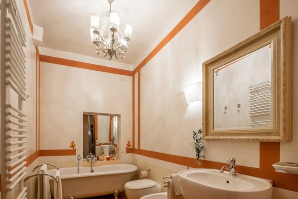 Une salle de bain avec deux lavabos, une baignoire et des toilettes. On y trouve un lustre, un miroir et un porte-serviettes.