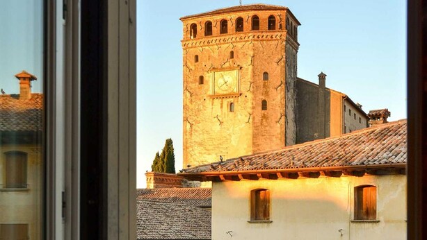 A view of a historic building with a tower and a flag, through a window with a balcony. - Palazzo Benassi Nobile (Asolo)