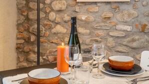 A dining table set with plates, bowls, glasses, and cutlery, a candle, and a bottle of wine, against a stone wall with a framed artwork above. - Green Lodge - Apartment 3 - 1 BR 1 Bath (Cressa)