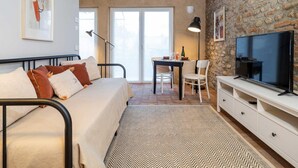 A cozy room with a bed, a dining table, chairs, a TV, and a stone wall. - Green Lodge - Apartment 3 - 1 BR 1 Bath (Cressa)