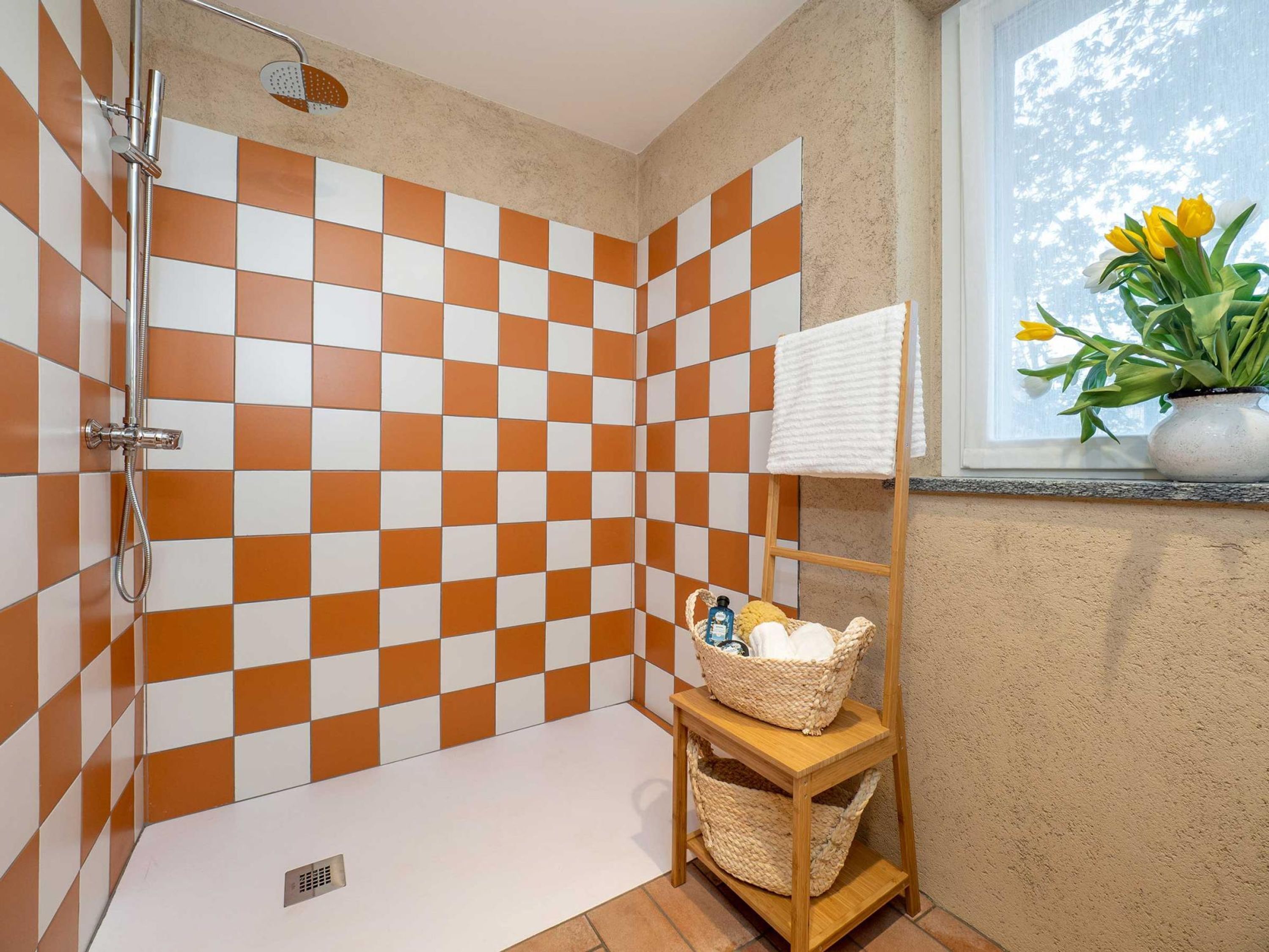 A bathroom with checkered orange and white tiles, a shower, a wooden shelf with baskets, and a window with a view of trees and flowers.