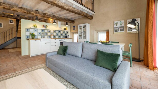 A spacious living area with a grey sofa, green cushions, a patterned rug, and a dining table with chairs. A kitchen with white cabinets and blue flower tiles is visible in the background. - Green Lodge - Apartment 5 - 1 BR 1 Bath (Cressa)